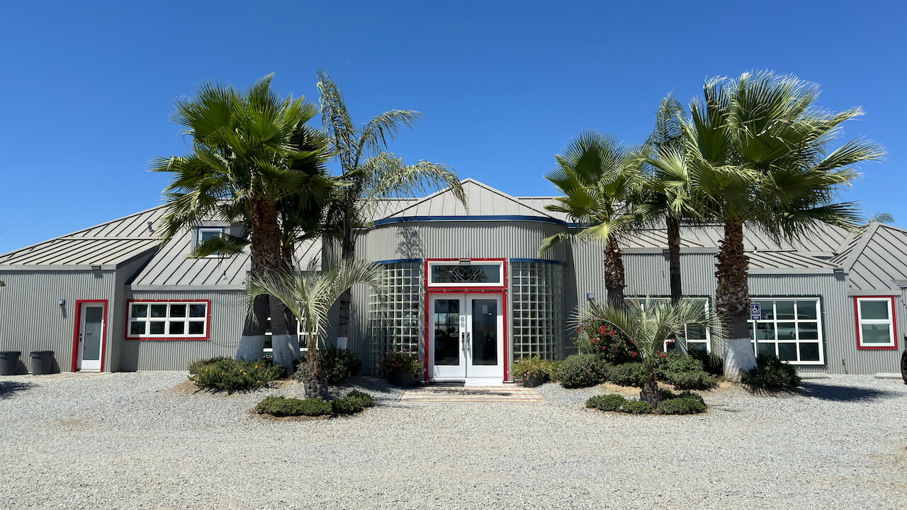 A single-story, modern gray building with red and white accents and palm trees on either side of the doorway.