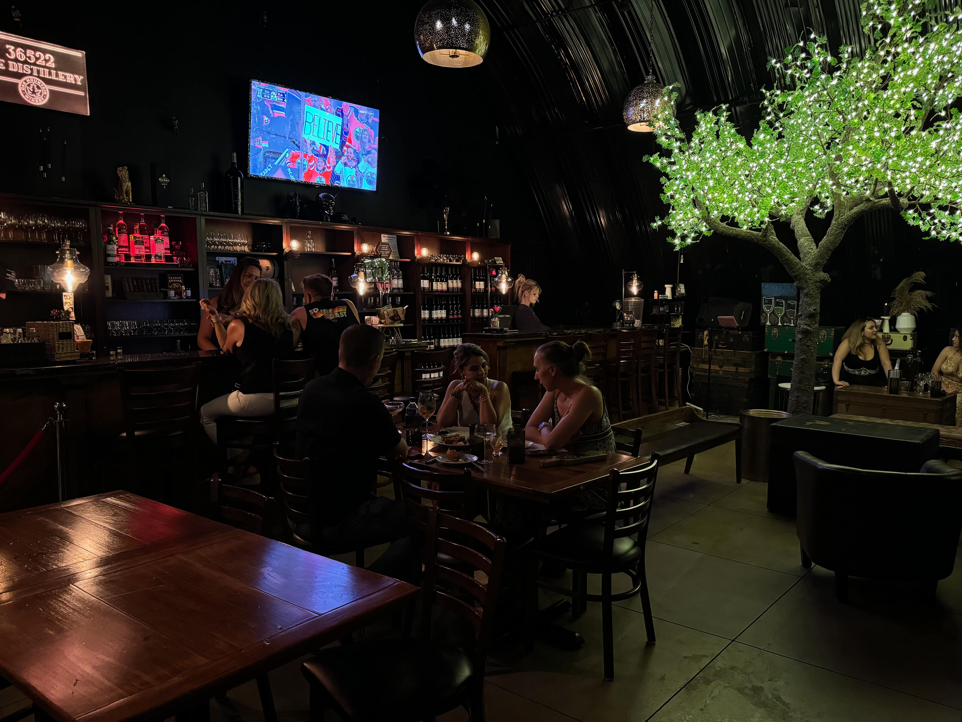A dark bar with tvs, people at a table, and tree with green lights for leaves.